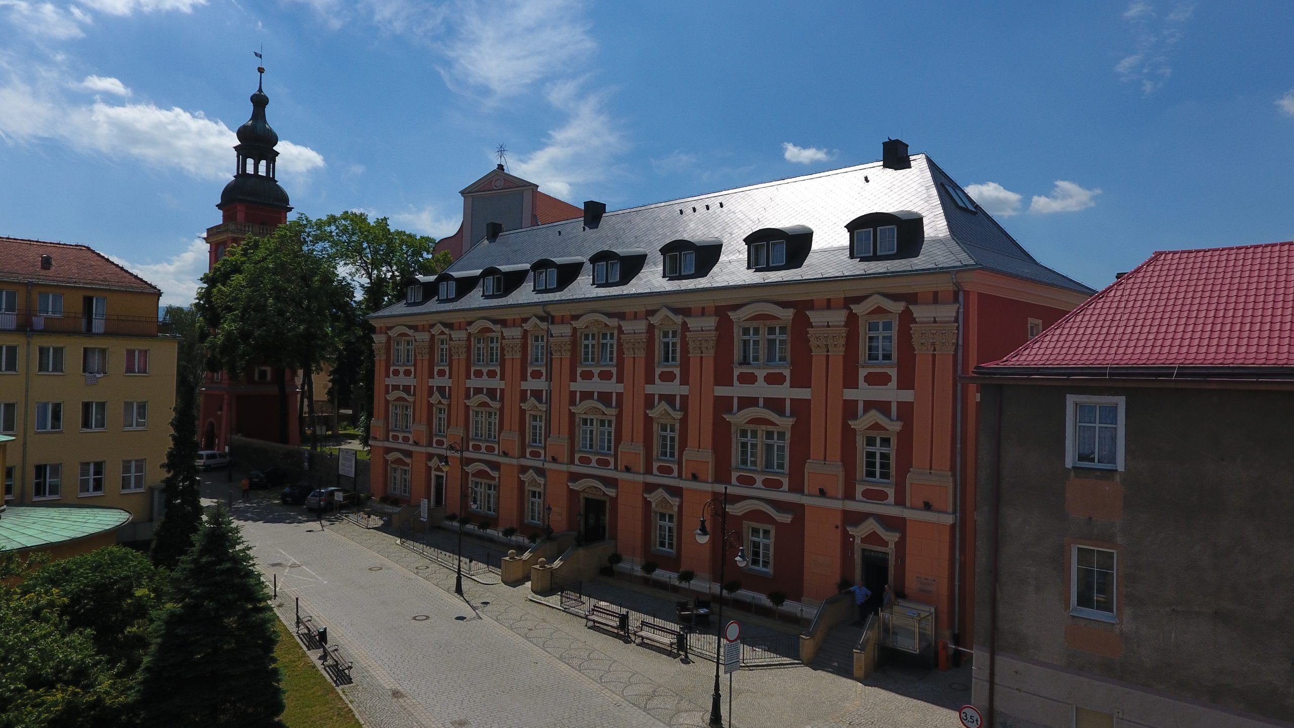 Długi, trójkondygnacyjny barokowy budynek, pięknie zrewitalizowany, pokryty kolorami w odcieniach pomarańczy i czerwieni. Na drugim planie wieża kościelna.