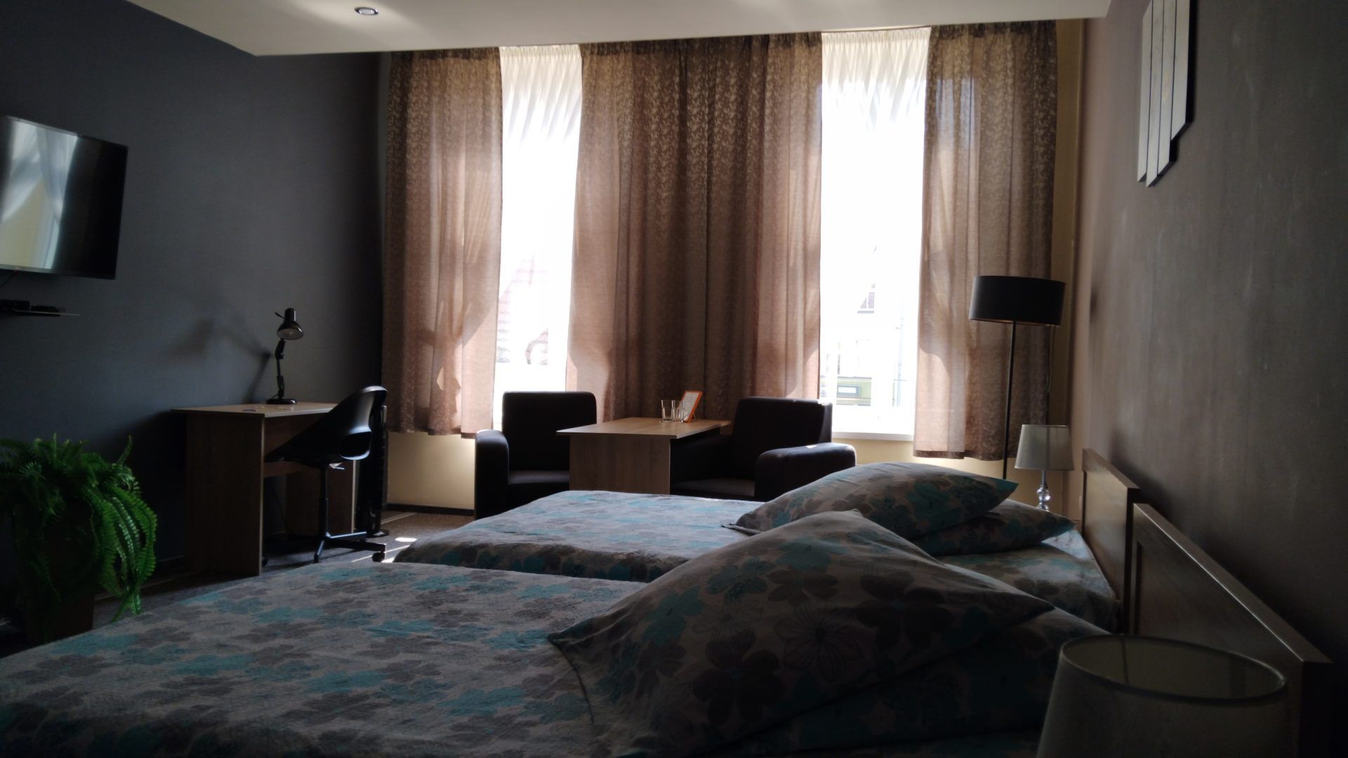 Przestronny pokój hotelowy z dwoma łóżkami pojedynczymi, biurkiem do pracy i wygodnym kącikiem wypoczynkowym przy oknie. Wnętrze utrzymane jest w stonowanych kolorach, z dużą ilością naturalnego światła wpadającego przez zasłonięte firany.