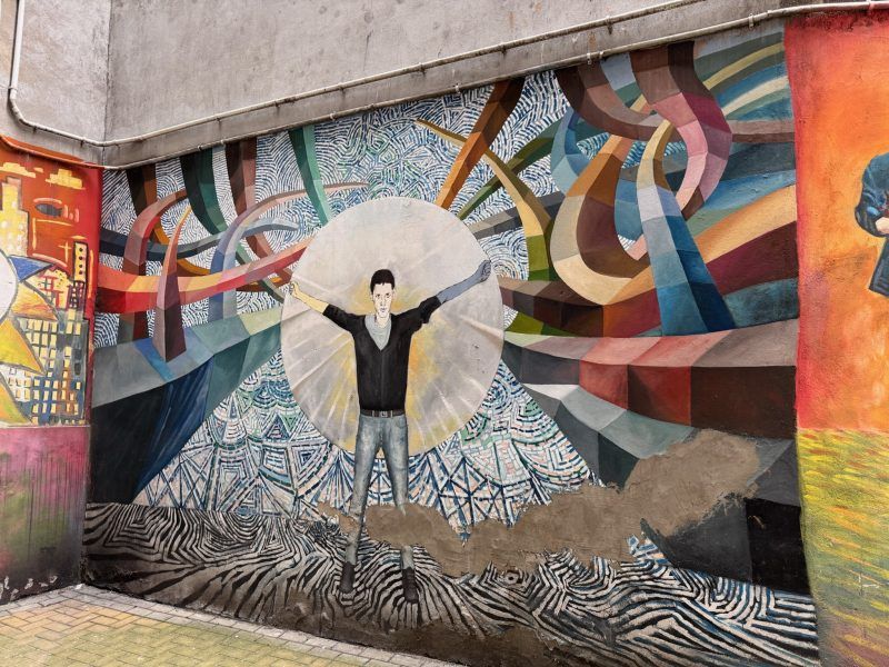 Zdjęcie przedstawia kolorowe podwórka i mural z chłopcem, który ma rozłożone ręce i jest otoczony przez abstrakcyjne figury i linie.