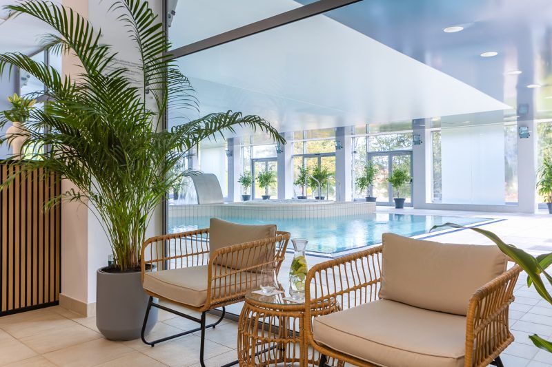 Nowoczesna strefa rekreacyjna z przeszklonymi ścianami oferuje kryty basen z atrakcjami wodnymi oraz komfortową przestrzeń do relaksu. Wygodne fotele i roślinność tworzą przyjazną atmosferę sprzyjającą odpoczynkowi.