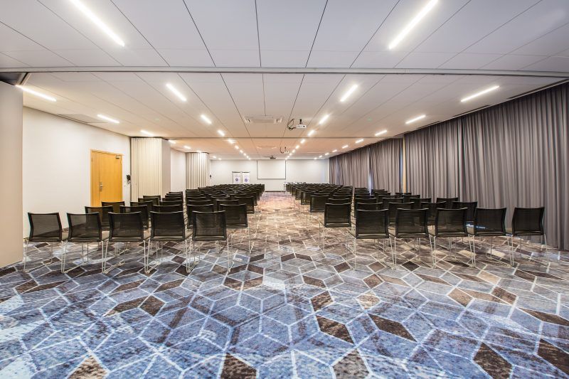 Duża sala konferencyjna z wieloma rzędami pojedynczych, czarnych krzeseł. 
