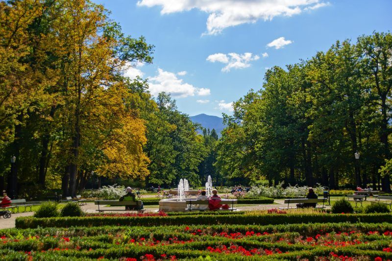 Piękny, zadbany park z równo przyciętą trawą i krzewami ozdobnymi. Na dalszym planie fontanna oraz górski krajobraz.
