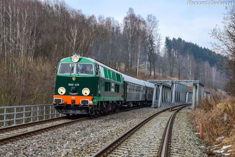 Zdjęcie przedstawia widok na dwa tory i pociąg jadący po jednym z nich na tle jesiennej pogody i niewielkich wzniesień