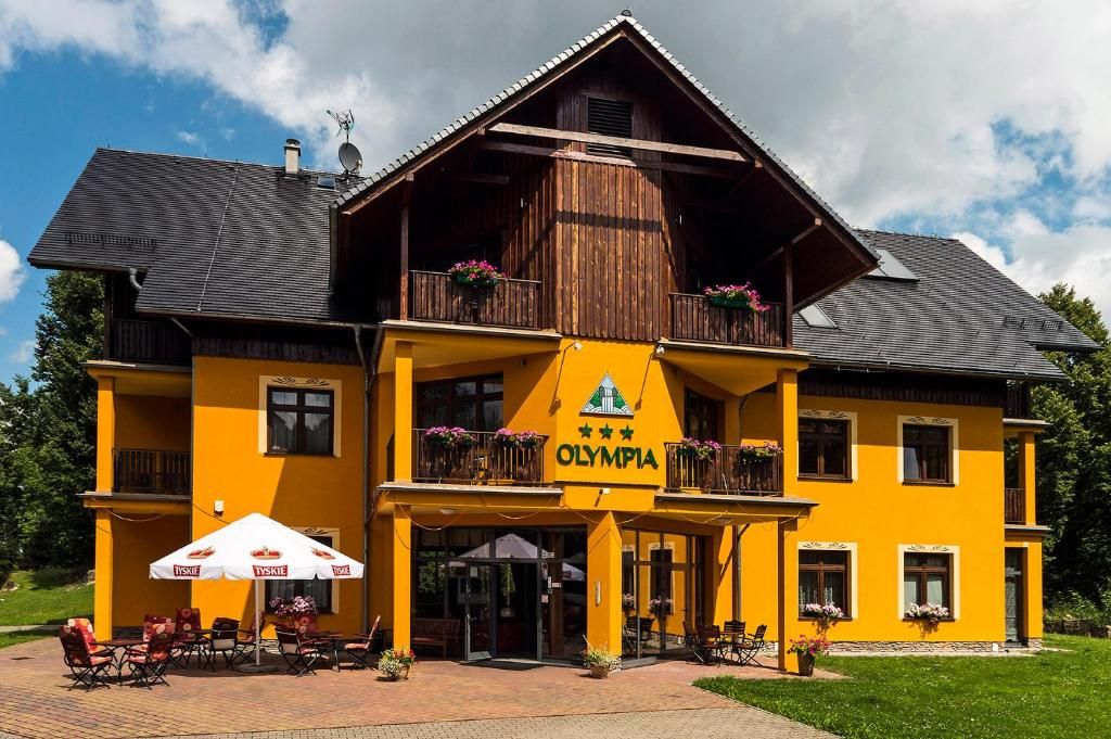 Urokliwy budynek hotelu Olympia z charakterystyczną pomarańczową elewacją i balkonami ozdobionymi kwiatami. Przed wejściem znajduje się taras z parasolami i stolikami, idealny do odpoczynku w słoneczne dni.