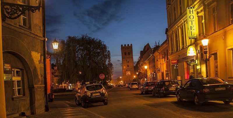 Wieczorny widok na ulicę w centrum miasta z charakterystyczną wieżą w tle. Po prawej stronie widać hotel z jasno podświetlonym szyldem, a po lewej staromiejską zabudowę.