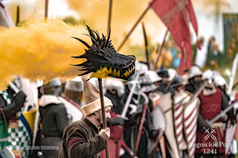 Głowa smoka zamieszczona na długim kiju, który trzyma mężczyzna w stroju z epoki.