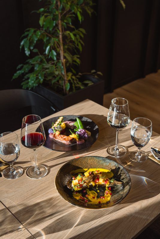 Na zdjęciu widać elegancko nakryty stolik w restauracji. Na stole ustawiono dwa wykwintnie podane dania oraz kieliszki z czerwonym winem i wodą. W tle widoczna jest doniczka z zieloną rośliną, a całość tworzy przytulną, nowoczesną atmosferę.
