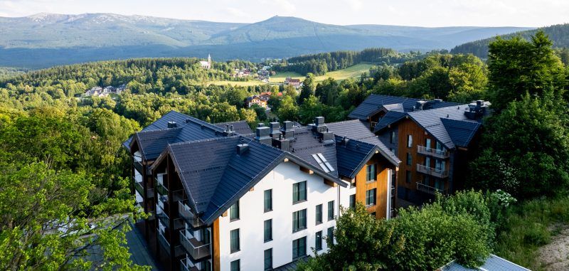 Malownicza panorama Szklarskiej Poręby z widokiem na pasmo Karkonoszy oraz kompleks budynków Happy Valley Resort wkomponowany w zielone otoczenie. W tle widać Śnieżne Kotły oraz charakterystyczną górę Szrenicę, co podkreśla górski, wypoczynkowy charakter miejsca.