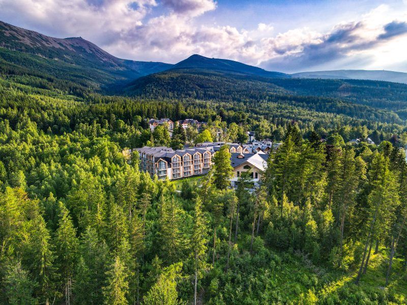 Na zdjęciu widzimy luksusowy kompleks Green Mountain Hotel & Apartments, malowniczo położony pośród gęstych lasów u podnóża Karkonoszy, z majestatyczną Śnieżką górującą w tle. Obiekt harmonijnie wtapia się w górski krajobraz, oferując spokój, bliskość natury i spektakularne widoki.