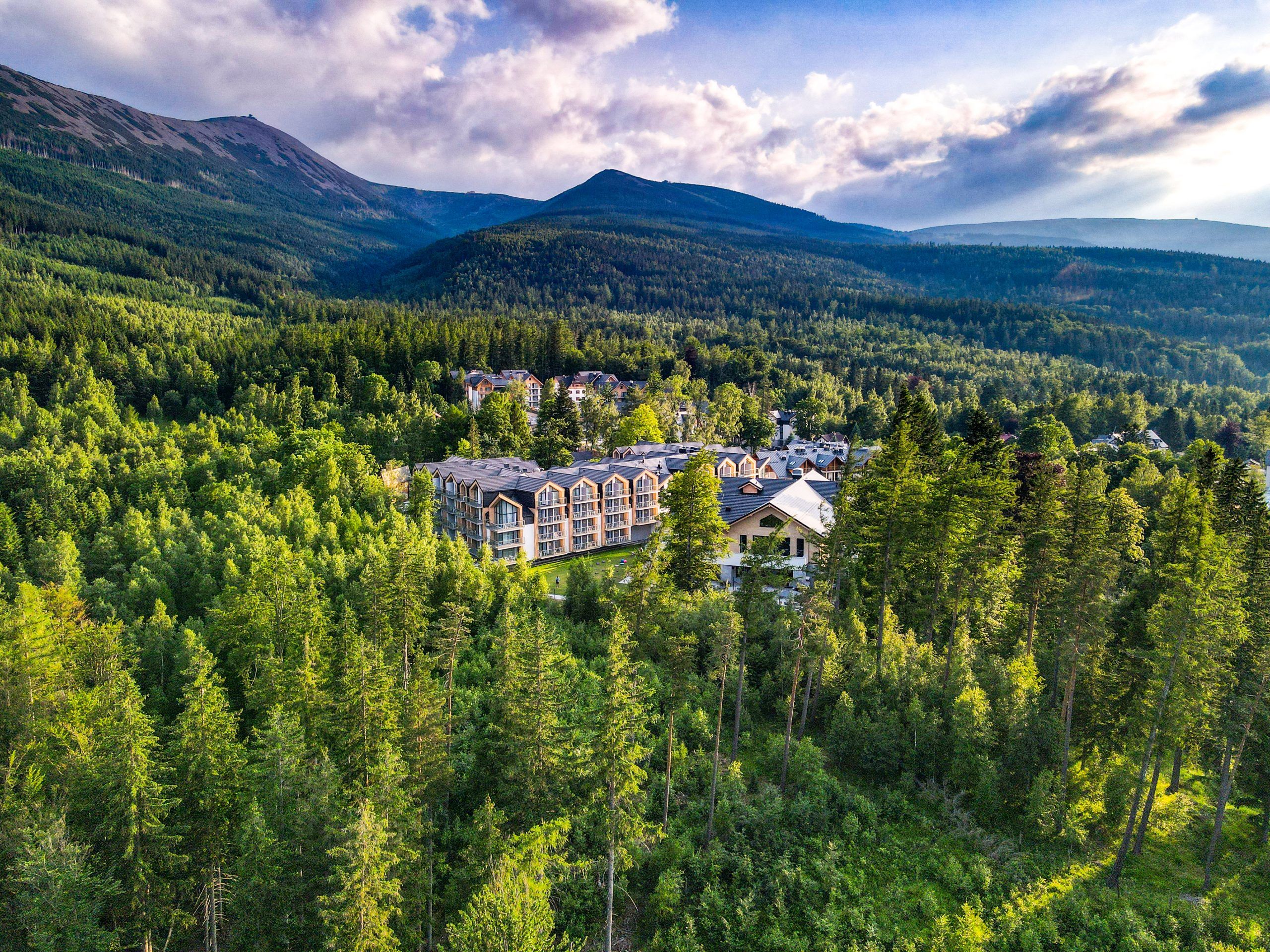 Na zdjęciu widzimy luksusowy kompleks Green Mountain Hotel & Apartments, malowniczo położony pośród gęstych lasów u podnóża Karkonoszy, z majestatyczną Śnieżką górującą w tle. Obiekt harmonijnie wtapia się w górski krajobraz, oferując spokój, bliskość natury i spektakularne widoki.