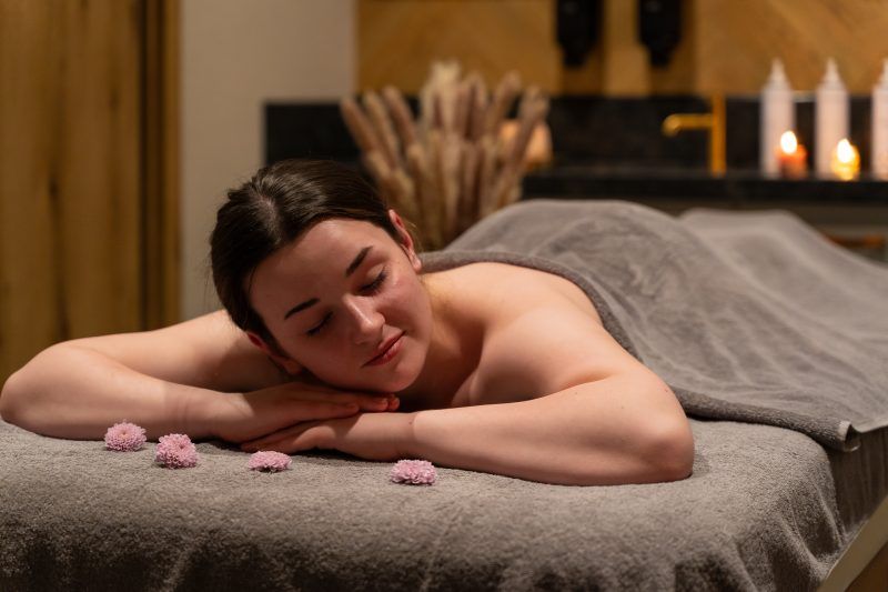 Kobieta podczas relaksacyjnego masażu w eleganckim wnętrzu spa, gdzie panuje przytulna i spokojna atmosfera. Subtelne światło, świeczki i kwiaty na stole potęgują wrażenie komfortu i odprężenia.