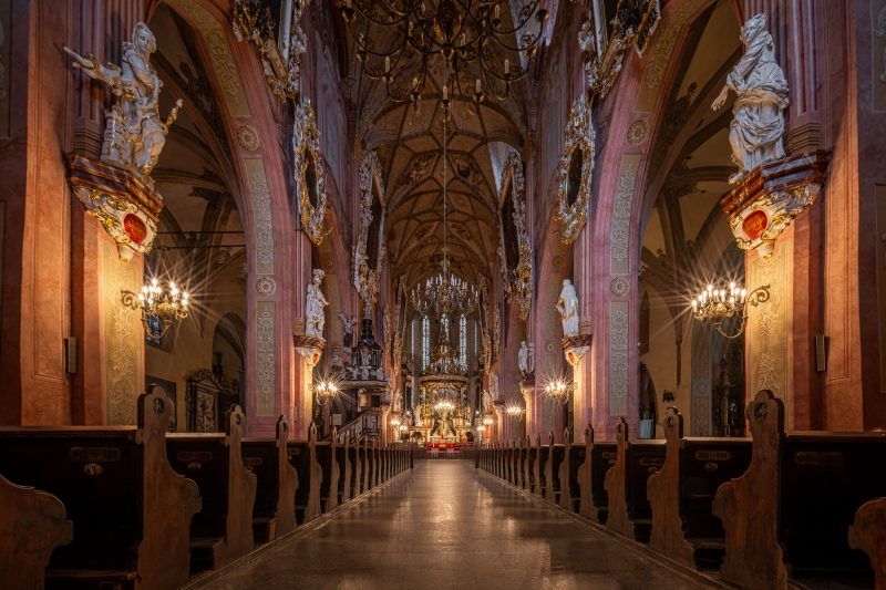 Przestronna nawa główna, pełna światła wpadającego przez witraże, z bogato zdobionymi sklepieniami i wysokimi filarami, które prowadzą wzrok ku wielkim ołtarzom i rzeźbom.