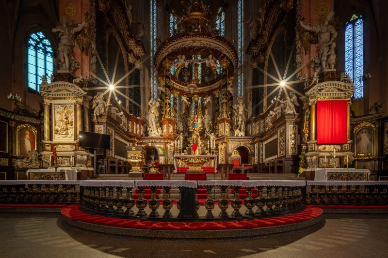 Przestronna nawa główna, pełna światła wpadającego przez witraże, z bogato zdobionymi sklepieniami i wysokimi filarami, które prowadzą wzrok ku wielkim ołtarzom i rzeźbom.