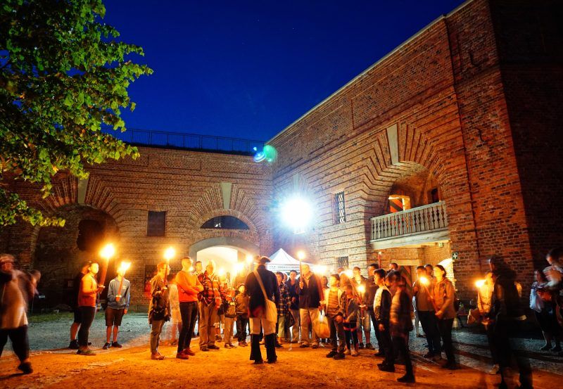 Grupa turystów z lampionami zgromadzona na dziedzińcu twierdzy
