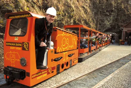 Il trenino turistico arancione della Miniera d'Oro di Złoty Stok attende i passeggeri davanti all'ingresso roccioso, con il macchinista in piedi accanto alla locomotiva.