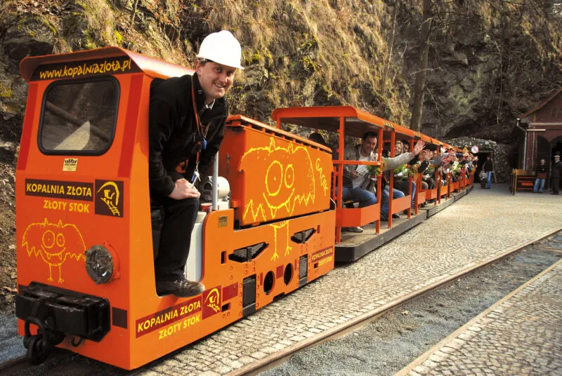 Il trenino turistico arancione della Miniera d'Oro di Złoty Stok attende i passeggeri davanti all'ingresso roccioso, con il macchinista in piedi accanto alla locomotiva.