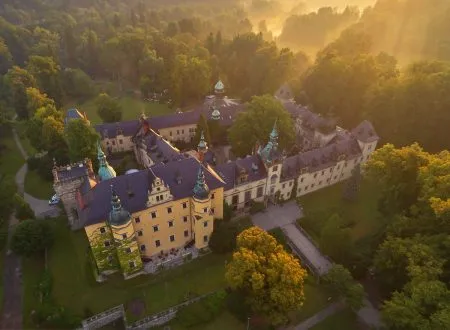 L'ampio Castello di Kliczków con le sue torri, il cortile e i tetti rossi è visto dall'alto tra fitti alberi illuminati dal sole caldo.